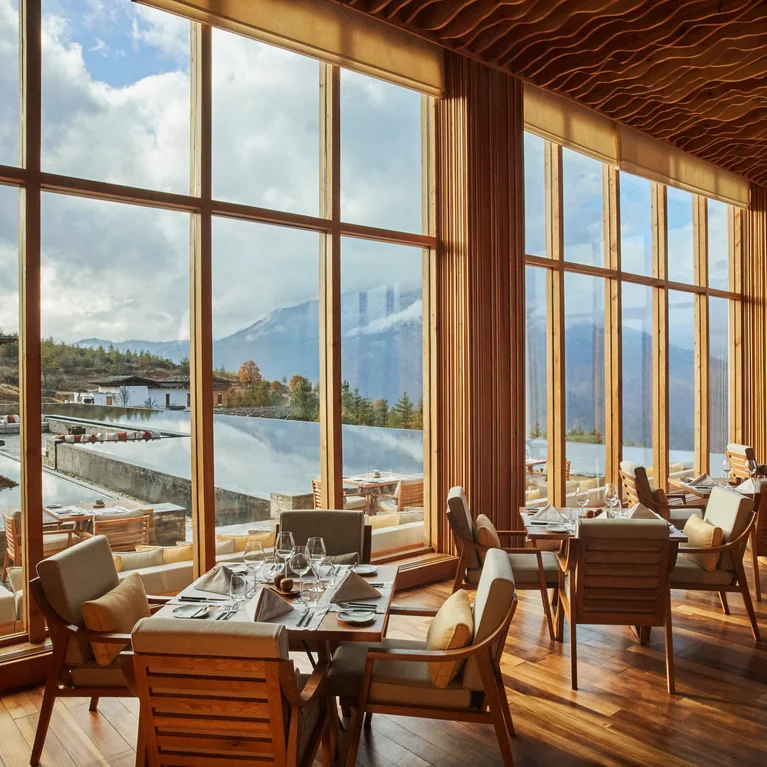 Restaurant with natural wood furnishings and floor-to-ceiling windows, offering views of mountains and a calm reflecting pool.