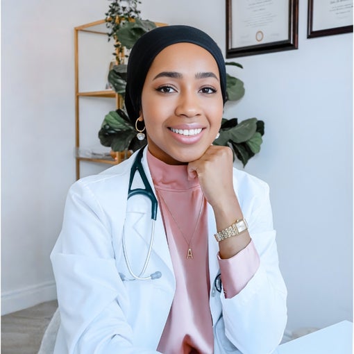 A woman in a white coat and headscarf sits at her desk, a stethoscope around her neck. Framed certificates hang on the wall behind her in a calm, professional space.