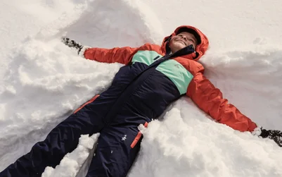 一个穿着橙色和藏青色连体雪服的孩子躺在雪地里，正摆出雪天使的姿势。.