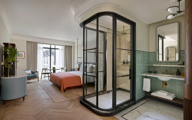 Contemporary hotel room featuring a double bed with orange accent, patterned curtains, light blue door, blue armchair, TV on a blue cabinet, and modern decor elements.