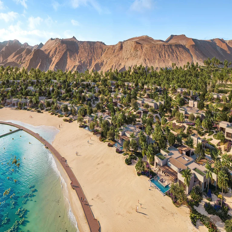 Villas and palm trees line a peaceful beach resort beside a tranquil lagoon, with mountains visible in the distance beneath a clear sky.