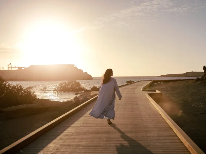 A guest enjoys a peaceful stroll along a wooden boardwalk beside calm coastal waters at sunset, framed by soft light and tranquil surroundings.