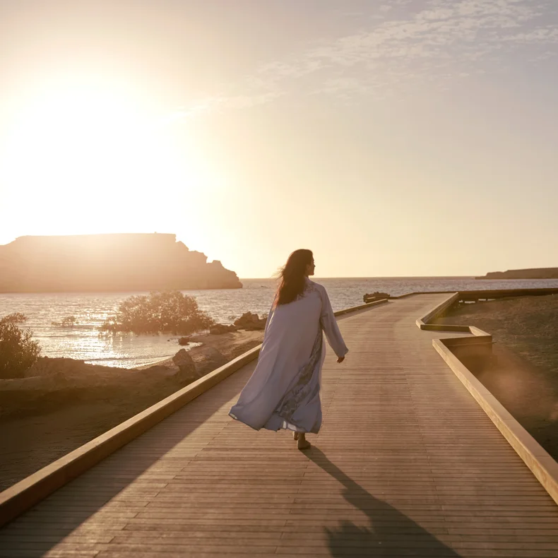 A guest enjoys a peaceful stroll along a wooden boardwalk beside calm coastal waters at sunset, framed by soft light and tranquil surroundings.