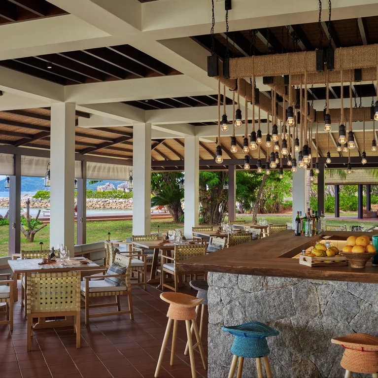 Restaurant space featuring natural wood tables and chairs, a stone bar with vibrant stools, soft pendant lights, and views of surrounding greenery and ocean.