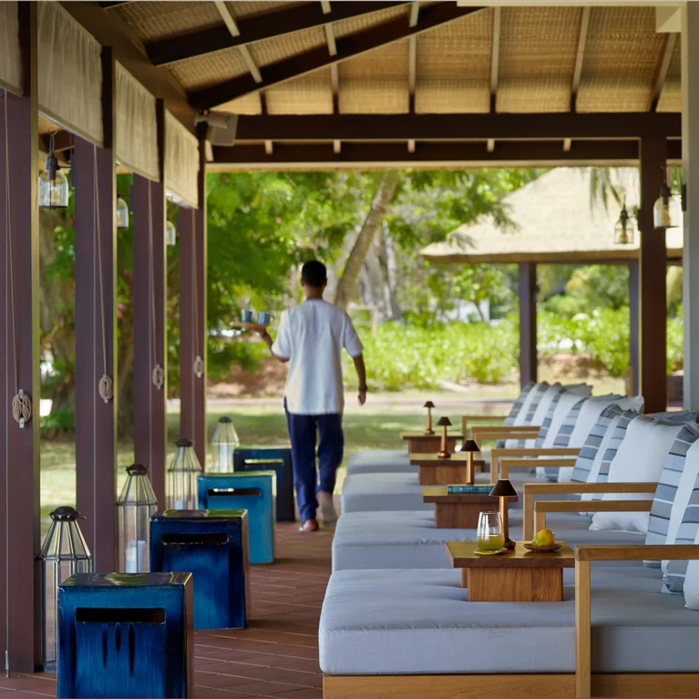 A guest strolls along a shaded veranda with cushioned chairs, small tables, and lanterns, surrounded by greenery in a tropical garden setting.