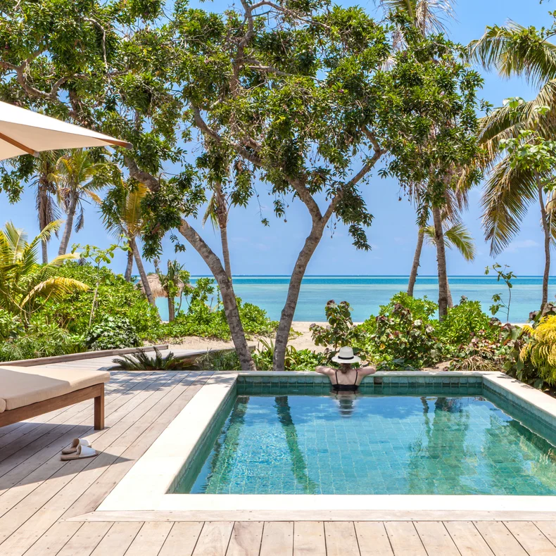 Outdoor setting with a serene pool, wooden deck, sun lounger, umbrella, surrounding greenery, and an open view toward the sand and clear blue sea.