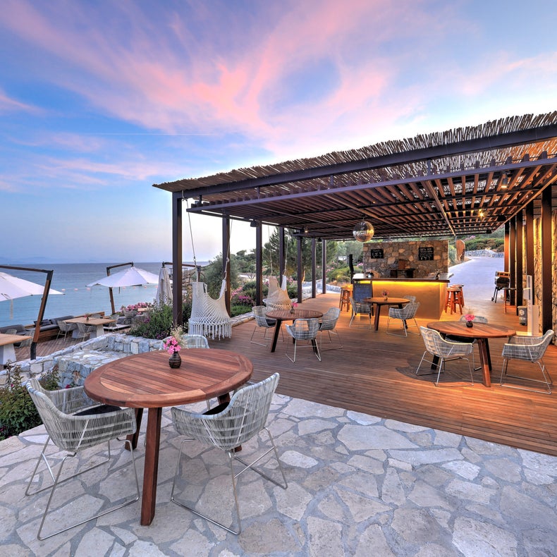 Un restaurante frente al mar con terrazas de madera, mesas y sillas modernas, un bar y vistas a la puesta de sol sobre las tranquilas olas bajo un cielo azul rosado.