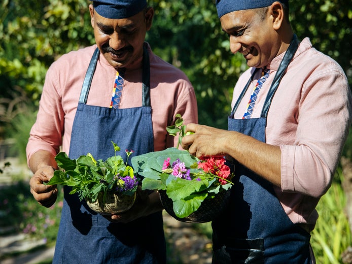 Unos cocineros con delantales y gorros azules sostienen cuencos con verduras y flores recién cosechadas en un jardín.