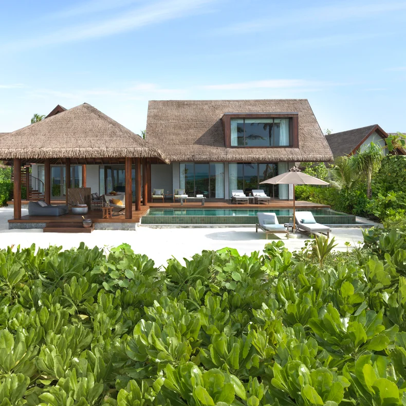 Villa with natural thatch roof, private pool, sun loungers, and garden plants, designed for privacy and well-being, set beneath a clear blue sky.
