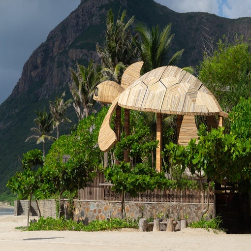Wooden sea turtle sculpture positioned above a beach hut, encircled by palm trees, greenery and a mountain landscape in the distance.