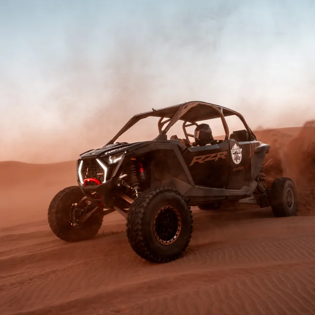 An off-road dune buggy moves across red desert sand, with light dust rising behind it under an open blue sky.