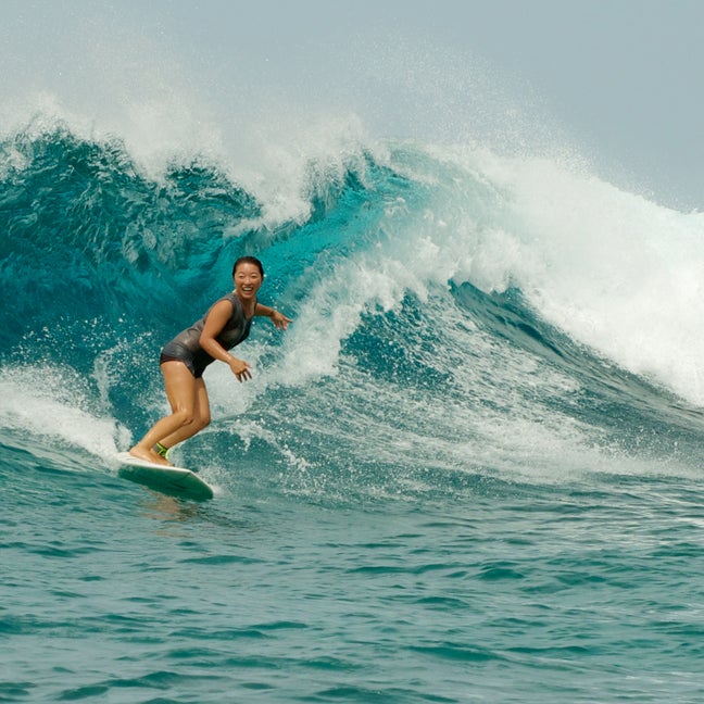 A guest enjoys surfing on a blue ocean wave, upright and balanced on a surfboard, capturing a moment of wellbeing in the natural surroundings.