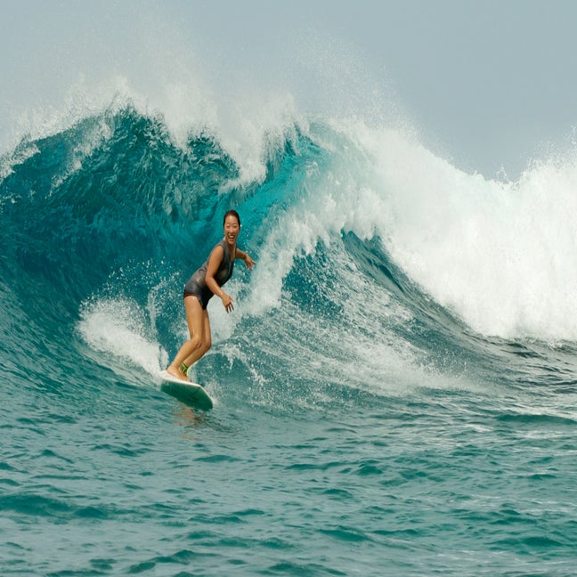 A guest enjoys surfing on a blue ocean wave, upright and balanced on a surfboard, capturing a moment of wellbeing in the natural surroundings.