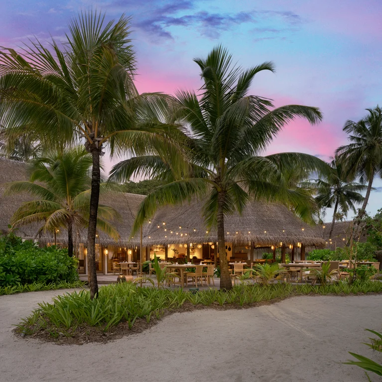 غرفة طعام ذات سقف طبيعي وأشجار نخيل وطاولات على أرضية رملية. تخلق سلاسل من الأضواء وهجاً ناعماً تحت سماء المساء الوردية والزرقاء.