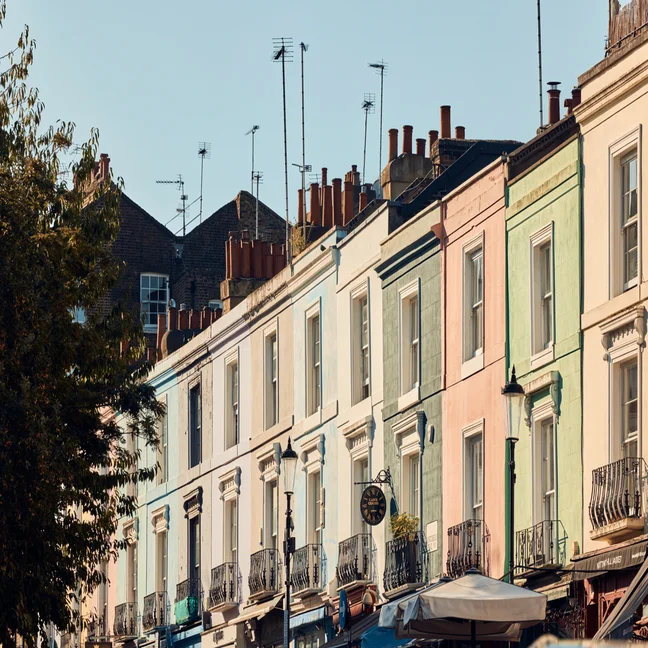 Pastel-hued terraced homes with balconies and chimneys stand beneath a clear sky, with leafy trees to the side, inviting a tranquil neighborhood setting.