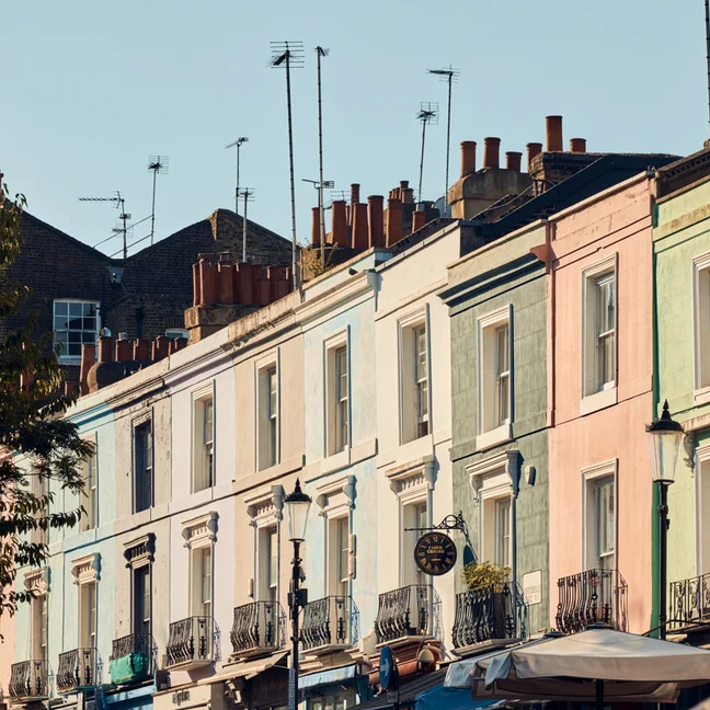 Pastel-hued terraced homes with balconies and chimneys stand beneath a clear sky, with leafy trees to the side, inviting a tranquil neighborhood setting.