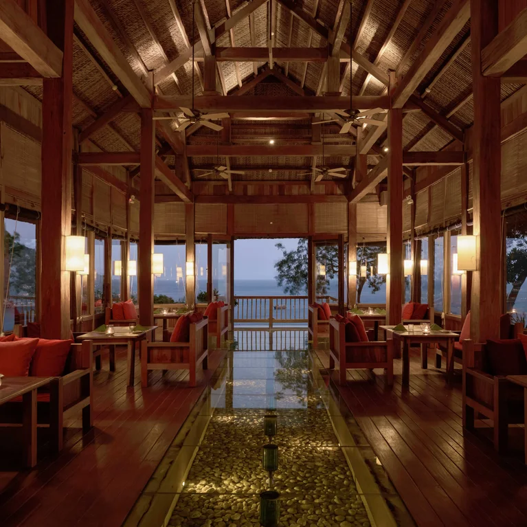 تتميز منطقة تناول الطعام في الهواء الطلق بسقف من الخشب الطبيعي، وإضاءة ناعمة، ومقاعد منجدة باللون الأحمر، وأرضية زجاجية على أرضية حجرية تطل على مناظر المحيط الرائعة عند غروب الشمس.