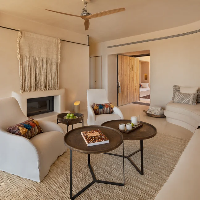 غرفة معيشة تتميز بألوان عضوية ناعمة وكرسيين بذراعين وأريكة منحنية وطاولات دائرية وعناصر منسوجة وتفاصيل من الخشب الطبيعي.