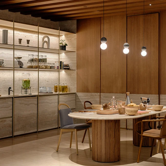Kitchen with natural wood and stone finishes, open shelves with curated objects and greenery, dining table set for a meal beneath pendant lights.
