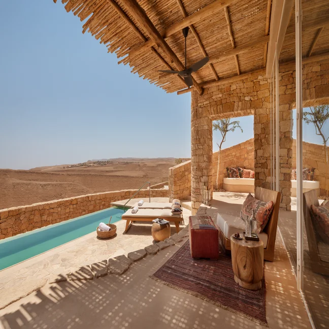 A terrace features a small pool, lounge seating, a rug, and wooden furniture beneath a pergola, overlooking an expansive desert landscape.