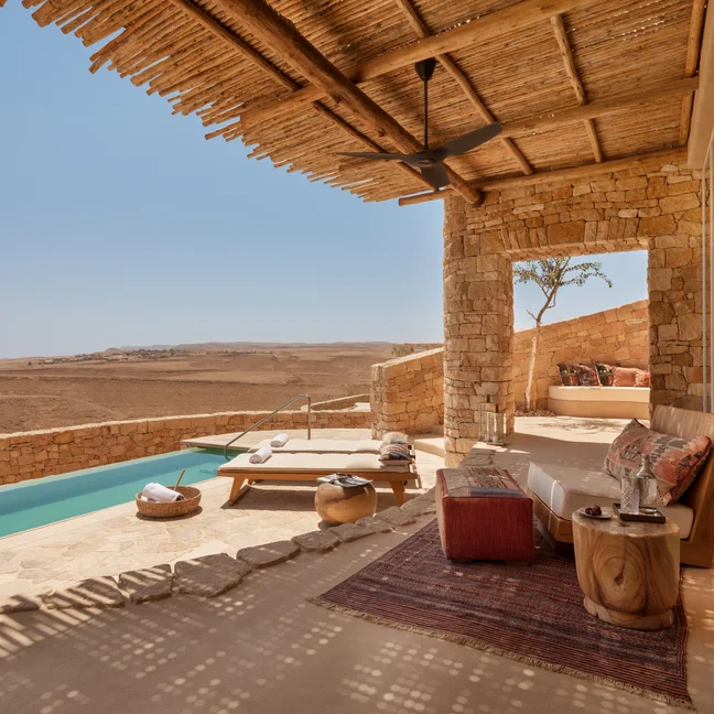 A terrace features a small pool, lounge seating, a rug, and wooden furniture beneath a pergola, overlooking an expansive desert landscape.