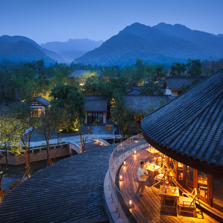Resort illuminated at dusk, set among trees and mountains. Guests dine on a curved outdoor terrace, connecting with nature in a tranquil setting.