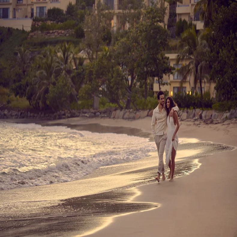 Two people walk barefoot along a sandy beach at sunset, gentle waves nearby and green foliage in the distance. The scene conveys tranquility and nature.