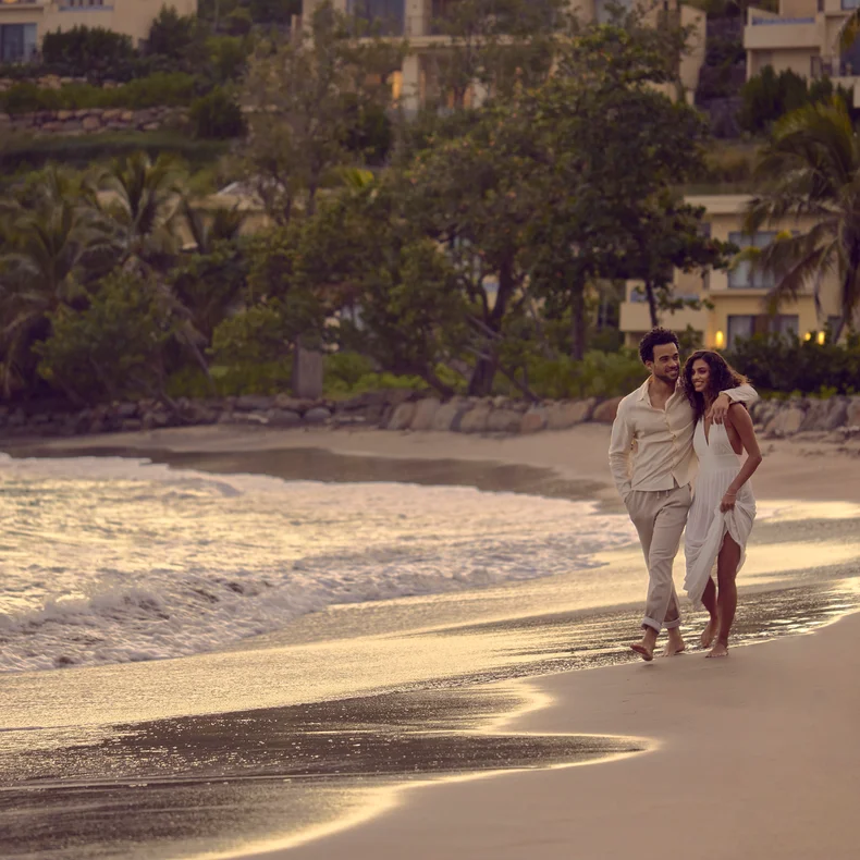Two people walk barefoot along a sandy beach at sunset, gentle waves nearby and green foliage in the distance. The scene conveys tranquility and nature.
