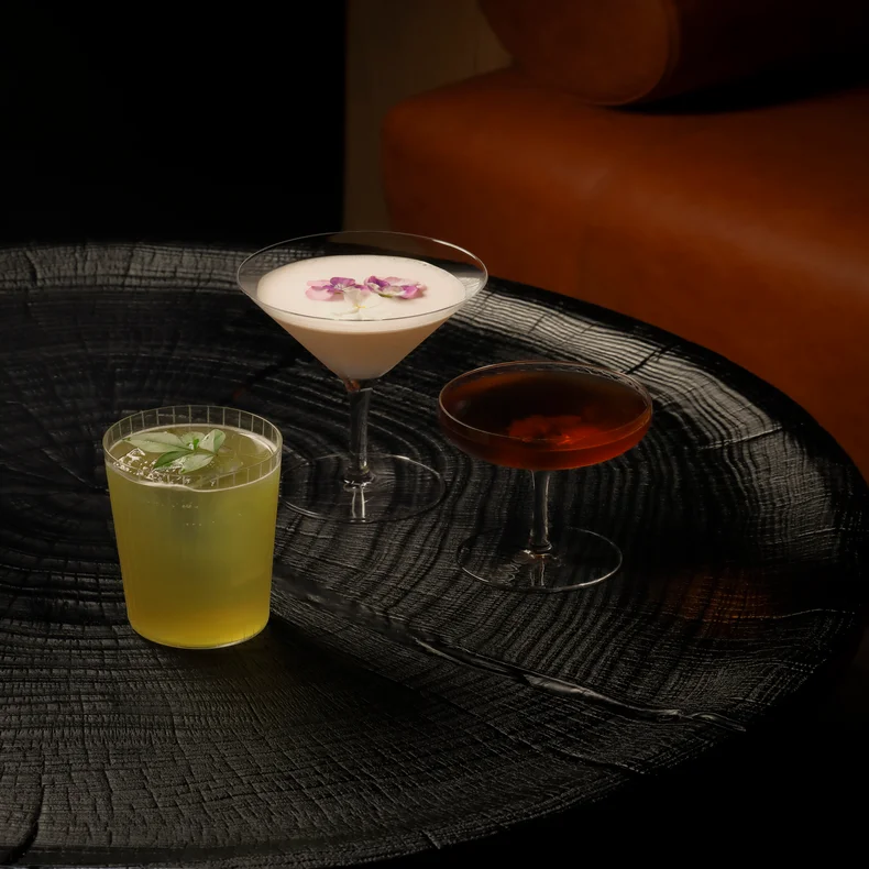 A selection of thoughtfully crafted beverages: a creamy white drink garnished with purple flowers, a refreshing yellow-green option with mint, and a rich amber drink in a coupe glass.