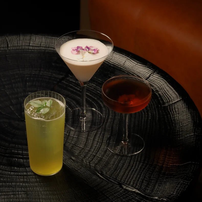 A selection of thoughtfully crafted beverages: a creamy white drink garnished with purple flowers, a refreshing yellow-green option with mint, and a rich amber drink in a coupe glass.