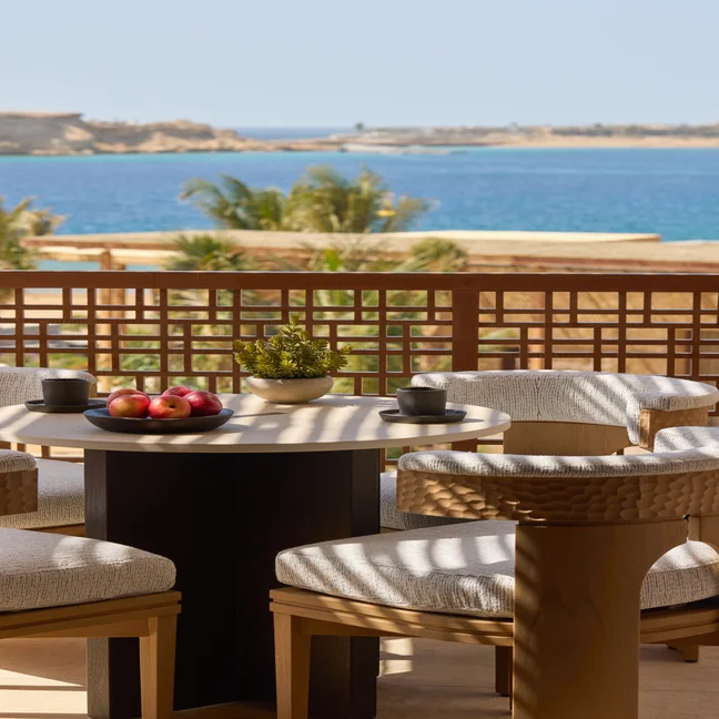 A terrace table set with apples, cups, and a potted plant is surrounded by four chairs, overlooking calm blue water and hills in the distance.