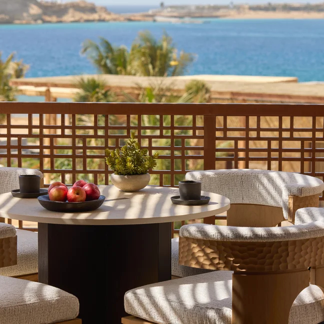 A terrace table set with apples, cups, and a potted plant is surrounded by four chairs, overlooking calm blue water and hills in the distance.
