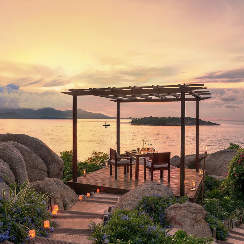 Outdoor dining table for two under a pergola on a wooden deck beside the ocean at dusk, with natural rock features and surrounding greenery.