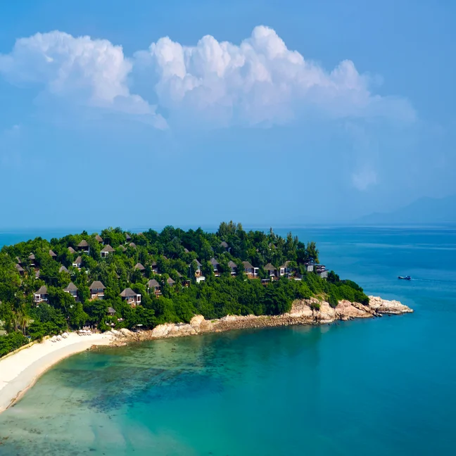 Green island with thoughtfully spaced villas, bordered by clear turquoise water and white sand beach, under a partly cloudy sky.