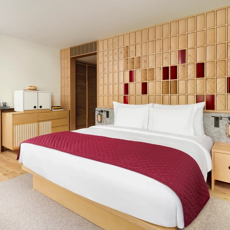 Hotel room featuring a king bed with organic white linens and a red quilt, light wood furnishings, and a wooden accent wall with red highlights behind the bed.