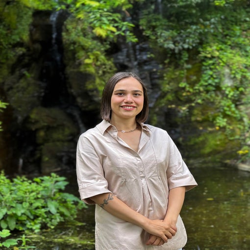 A guest in a light beige dress stands, smiling near green foliage and a gentle waterfall, reflecting a tranquil and natural setting.