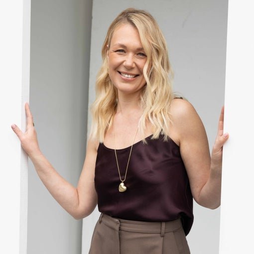 A woman with blonde wavy hair, in a dark sleeveless top, long necklace, and brown pants, stands in a doorway gently holding each side of the frame.