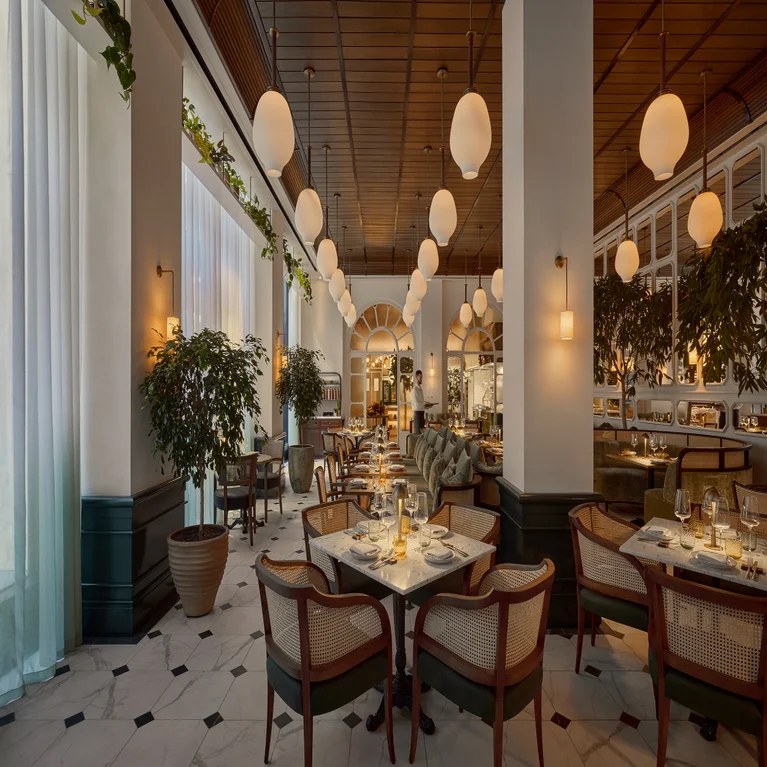 Restaurant with natural wood chairs, round dining tables, marble flooring, lush potted plants, gentle globe lighting, and tall windows with sheer curtains.