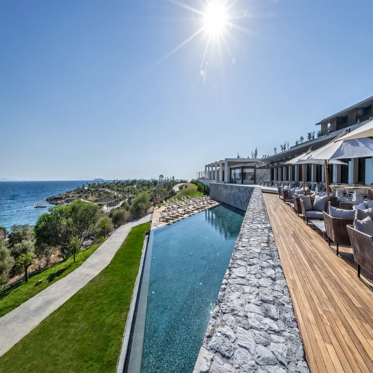 Infinity pool with sun loungers and shaded outdoor seating facing the ocean. Clear sky, blue water, and natural surroundings enhance relaxation and connection to nature.