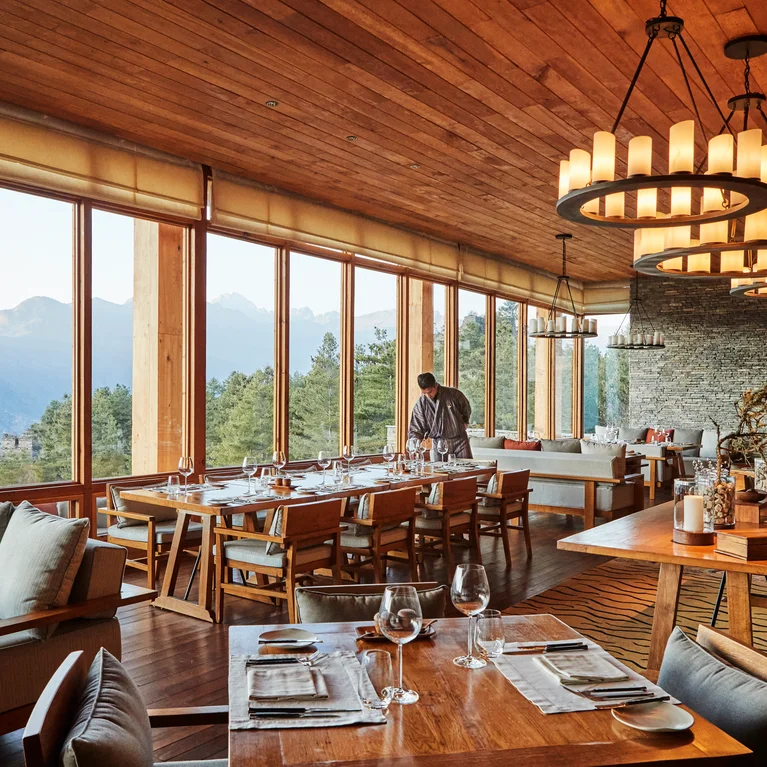 Restaurant with large windows framing mountain views, warm wood interiors, cushioned chairs, and organized dining tables. A person stands quietly near the windows.