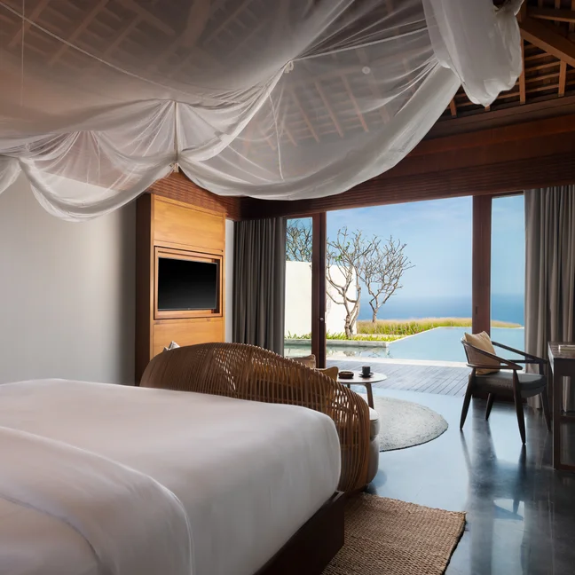 Guest room with white-canopied bed, wood furnishings, desk, and glass doors leading to a patio overlooking ocean and curated gardens.