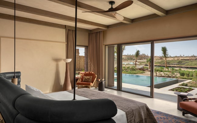 Spacious room featuring a king bed, leather chair, floor-to-ceiling windows with views of a private pool, garden with palm trees, and sunlit landscape.