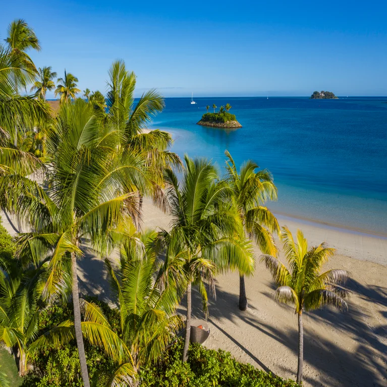 Expansive shoreline with soft sand, native palms, gentle blue sea, a distant island and sailboat beneath a sunlit sky.