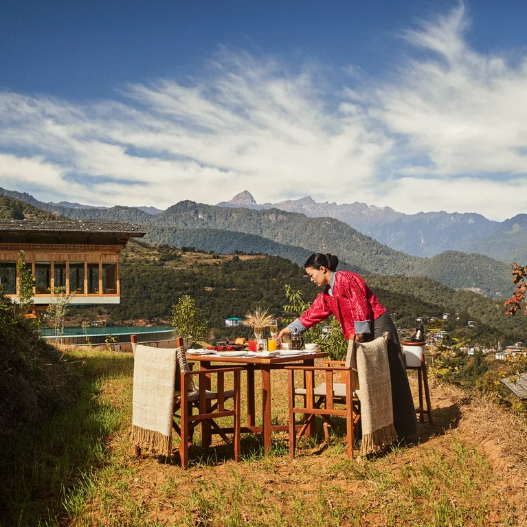 A person prepares a dining table outside on a grassy slope, overlooking mountains and clouds, next to a wooden building.