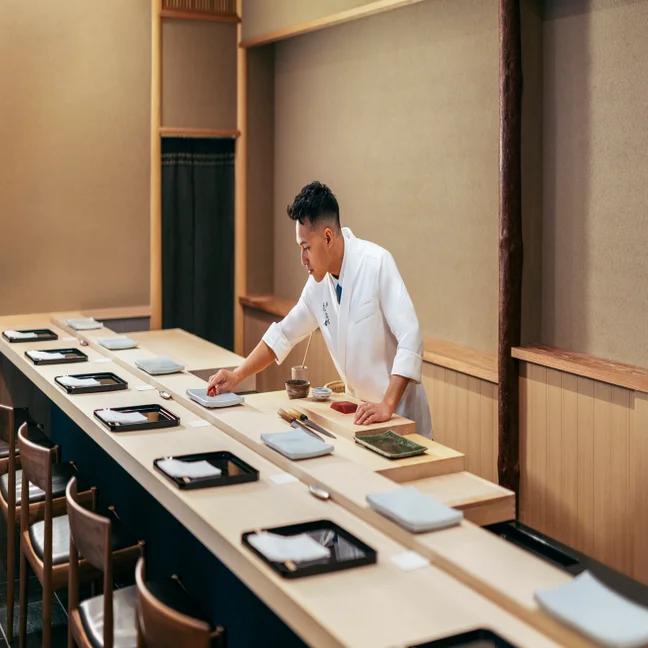 A culinary professional arranges fresh ingredients on a counter in a serene, contemporary dining space with thoughtfully set tables and natural lighting.