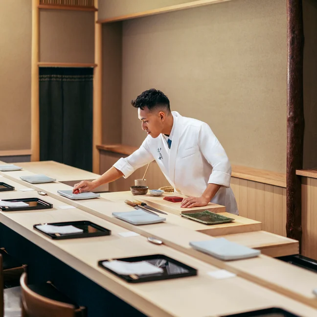 A culinary professional arranges fresh ingredients on a counter in a serene, contemporary dining space with thoughtfully set tables and natural lighting.
