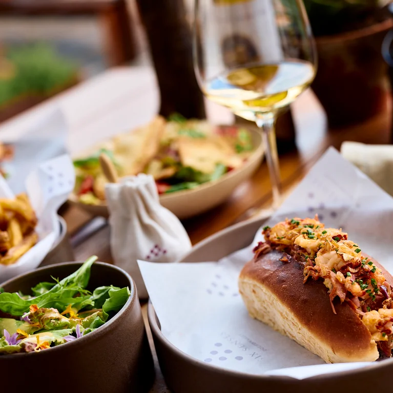 لفائف اللوبستر في كعكة طرية مع السلطة والبطاطا المقلية مع النبيذ الأبيض على طاولة خشبية؛ وتتميز الخلفية بأطباق إضافية غير واضحة.