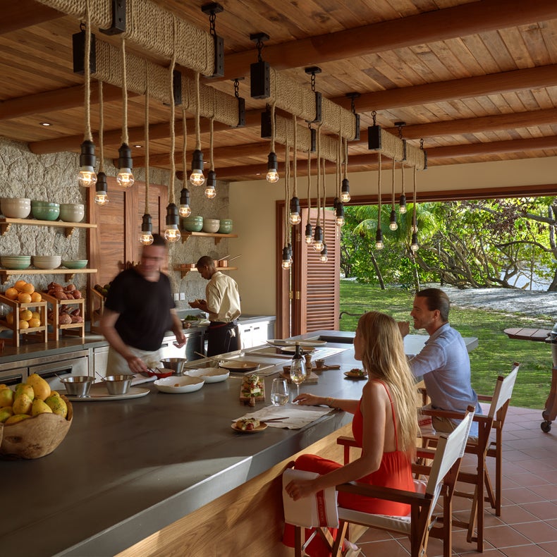 Cocina al aire libre donde los chefs preparan platos frescos para los comensales sentados en el mostrador, con vistas al jardín con árboles y agua de fondo.