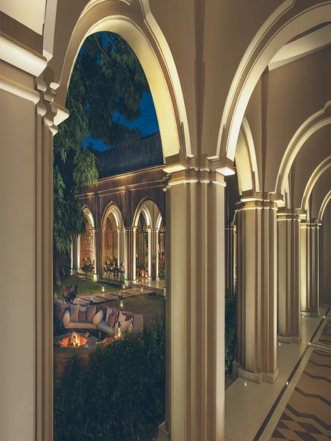 Una pasarela cubierta con columnas conduce a un patio suavemente iluminado por la noche, que ofrece asientos accesibles al aire libre, una exuberante vegetación y una suave iluminación.