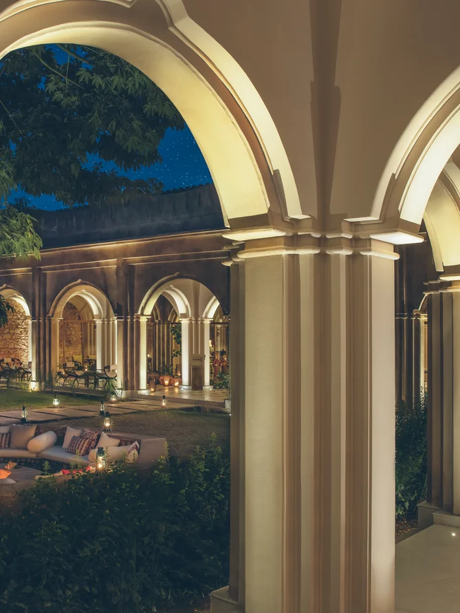 Una pasarela cubierta con columnas conduce a un patio suavemente iluminado por la noche, que ofrece asientos accesibles al aire libre, una exuberante vegetación y una suave iluminación.